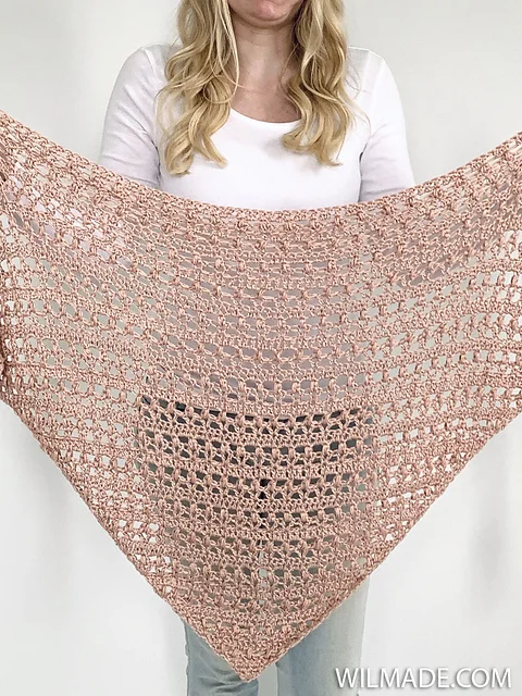 A woman holds up a large, triangular, light pink crocheted shawl with an open, lacy pattern—the perfect Vintage Tulip Shawl for adding timeless elegance to any outfit.