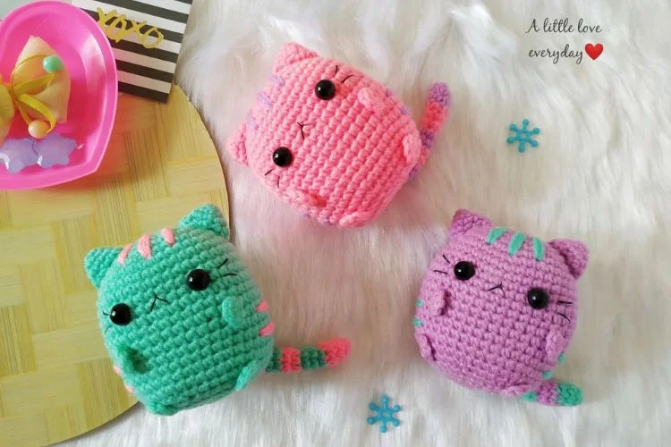 Three colorful crocheted cat toys on a fluffy white rug, with a pink bowl and decorative items nearby.