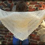 A person stands against a stone and brick wall, displaying a large, cream-colored Seems Like Old Times Shawl, crocheted in a triangular shape and elegantly draped across their back.