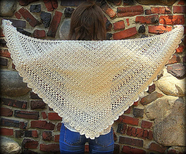 A person stands against a stone and brick wall, displaying a large, cream-colored Seems Like Old Times Shawl, crocheted in a triangular shape and elegantly draped across their back.
