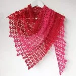 A triangular, lacy Hot Chili Shawl in shades of red and pink is draped over a white hanger against a plain white wall.