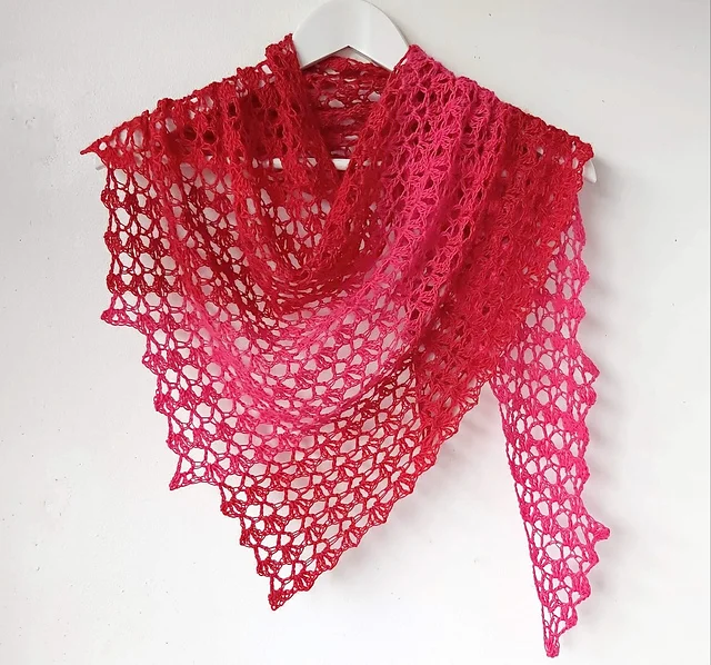 A triangular, lacy Hot Chili Shawl in shades of red and pink is draped over a white hanger against a plain white wall.