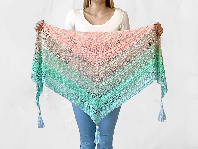 A person holds a Jaycee Butterfly Shawl, a triangular, lace-patterned piece featuring pink, green, and aqua gradient stripes with tassels at each corner.