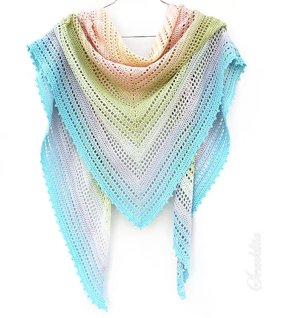 The Summer Morning Shawl is a triangular crochet piece with a lacy pattern in pastel gradient colors—peach, yellow, green, blue, and gray—hanging elegantly on a hanger against a white background.