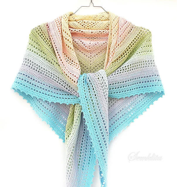 A crocheted triangular Summer Morning Shawl in pastel rainbow colors hangs on a hanger against a white background.