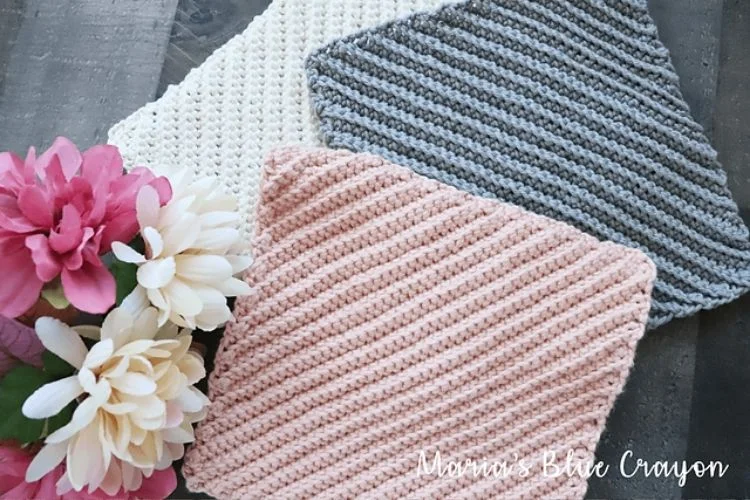 Assorted hand-knitted dishcloths in pastel tones arranged beside a bouquet of flowers.