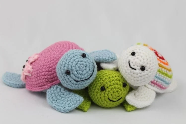 A group of three crocheted stuffed animals, including a pink turtle, a green turtle, and a white turtle with a colorful shell, smiling on a plain background.