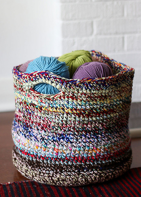 Multicolored Scrap Yarn Basket filled with blue, green, and purple yarn balls, placed on a wooden floor near a white brick wall.