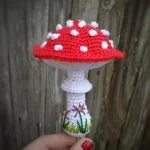 A hand holds a crocheted red and white mushroom—resembling classic toadstools amigurumi—with white spots and green grass embroidery on the stem, set against a wooden background.