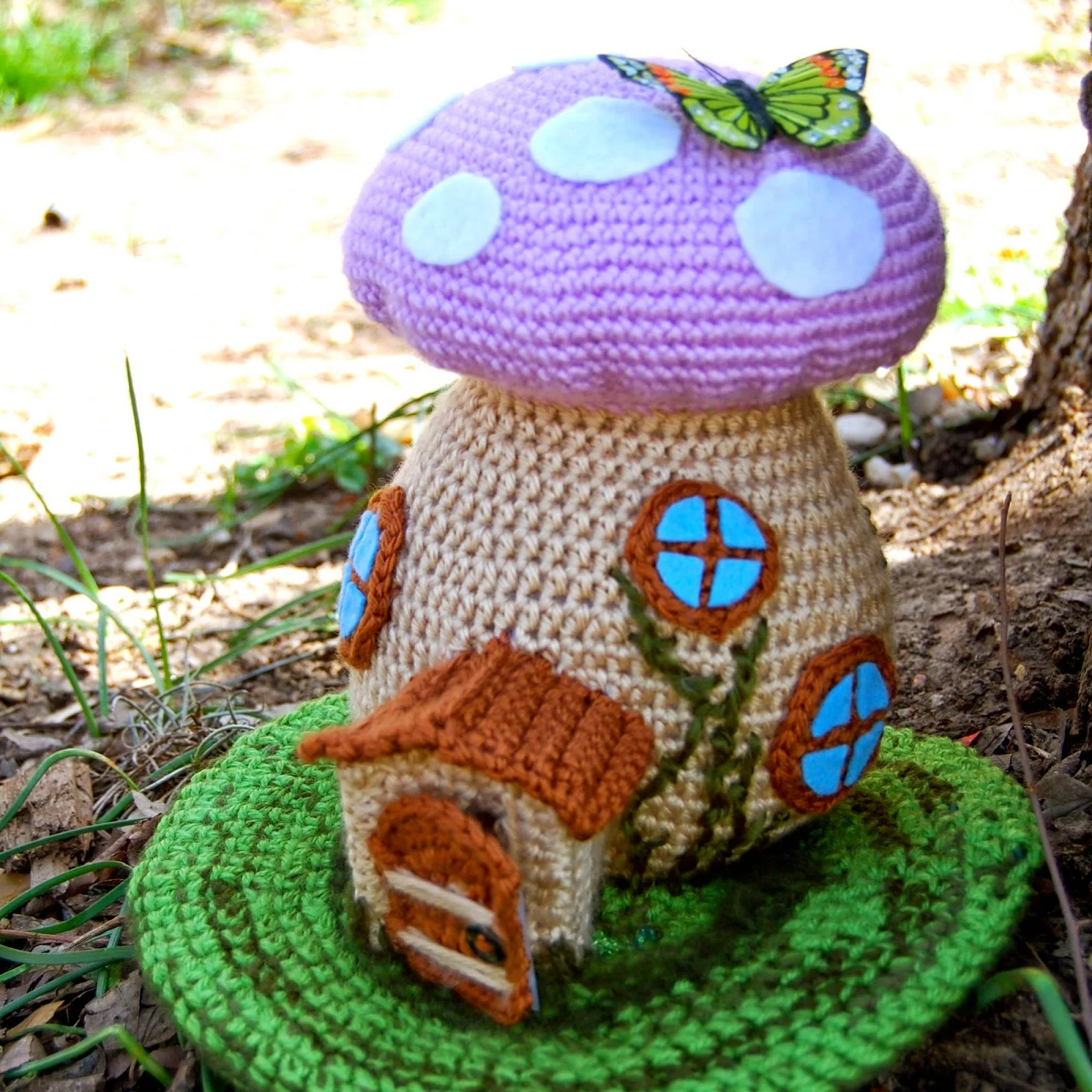 Crocheted Spring Fairy House Amigurumi with a pink cap, white spots, small door, blue windows, and a butterfly decoration, sitting on a green crocheted base outdoors.