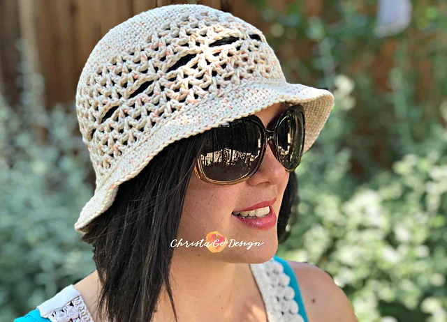 A woman wearing large sunglasses and a stylish Lunette Sun Hat outdoors, with greenery softly blurred in the background.