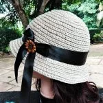 A beige crocheted hat, inspired by a Straw Sun Hat crochet pattern, with a wide black ribbon and decorative brooch is displayed on a mannequin head outdoors, surrounded by trees and greenery.