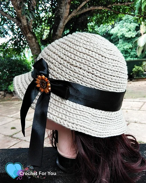 A beige crocheted hat, inspired by a Straw Sun Hat crochet pattern, with a wide black ribbon and decorative brooch is displayed on a mannequin head outdoors, surrounded by trees and greenery.