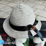 A beige straw sun hat crochet pattern with a black ribbon and bow, featuring a small brown flower decoration, rests on a black and white fence.
