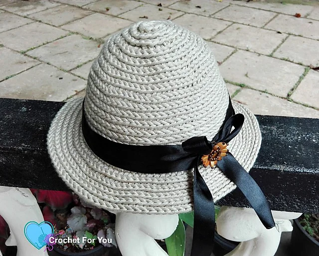 A beige straw sun hat crochet pattern with a black ribbon and bow, featuring a small brown flower decoration, rests on a black and white fence.
