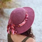 A person with curly hair wears a purple woven Desert Sun Hat adorned with a floral ribbon and bow, viewed from behind, outdoors.
