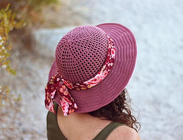 A person with curly hair wears a purple woven Desert Sun Hat adorned with a floral ribbon and bow, viewed from behind, outdoors.