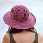 A person with curly hair, wearing a sleeveless top and a pink woven Desert Sun Hat, is seen from behind against a light background.