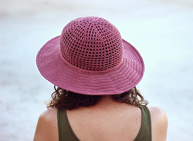 A person with curly hair, wearing a sleeveless top and a pink woven Desert Sun Hat, is seen from behind against a light background.