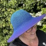 Person wearing a large, gradient sun hat in shades of blue and purple stands outdoors in front of green leafy bushes with small white flowers.