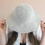 Person holding a June Sun Hat over their face, wearing a white shirt with black polka dots and a black top underneath.