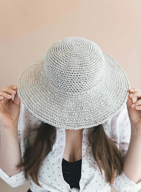 Person holding a June Sun Hat over their face, wearing a white shirt with black polka dots and a black top underneath.