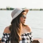 Woman wearing a June Sun Hat, sunglasses, and an off-shoulder plaid dress stands outdoors by a body of water, looking to the side.