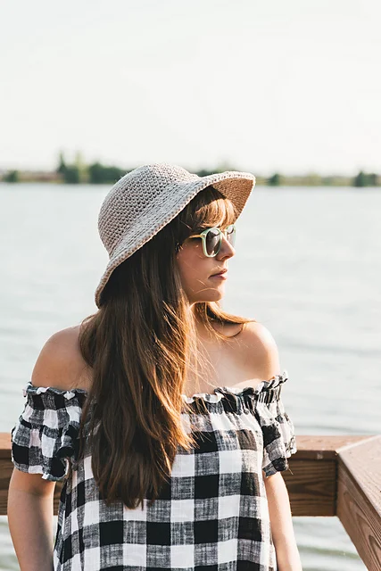 Woman wearing a June Sun Hat, sunglasses, and an off-shoulder plaid dress stands outdoors by a body of water, looking to the side.