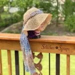 A mannequin head with purple hair wears the Stepping Out Sun Hat—a wide-brimmed crocheted hat—and a long, lacy scarf, displayed on a wooden railing that reads 