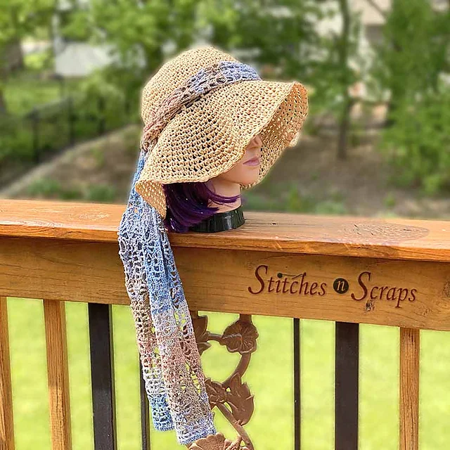 A mannequin head with purple hair wears the Stepping Out Sun Hat—a wide-brimmed crocheted hat—and a long, lacy scarf, displayed on a wooden railing that reads 
