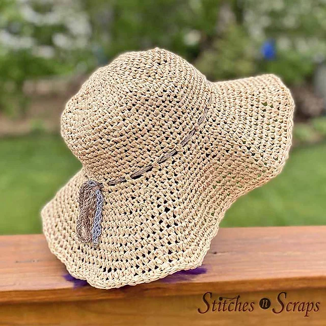 The Stepping Out Sun Hat is a wide-brimmed, crocheted straw hat with a simple brown accent, beautifully displayed on a wooden surface outdoors.