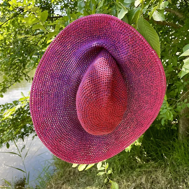 A pink and purple gradient sun hat hangs on a tree branch with green leaves near a body of water.
