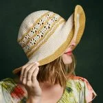 A woman with shoulder-length hair wears the Shangri-la Sunhat, a wide-brimmed woven hat with yellow and cream stripes, partially covering her face, against a dark green background.