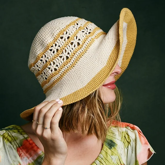 A woman with shoulder-length hair wears the Shangri-la Sunhat, a wide-brimmed woven hat with yellow and cream stripes, partially covering her face, against a dark green background.