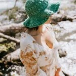 A person in a floral dress and a Summer Lovin' Crochet Sun Hat stands near a sunlit river, with blurred water and rocks in the background.