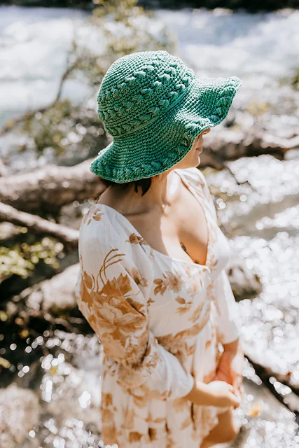 A person in a floral dress and a Summer Lovin' Crochet Sun Hat stands near a sunlit river, with blurred water and rocks in the background.
