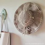 A Summer Lovin' Crochet Sun Hat with a tied bow hangs on a wall next to a white handbag.