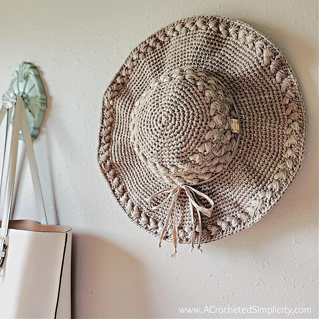 A Summer Lovin' Crochet Sun Hat with a tied bow hangs on a wall next to a white handbag.