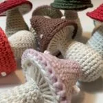 A group of Mushroom Amigurumi in various colors, including red, brown, pink, and green, are displayed closely together on a white surface.