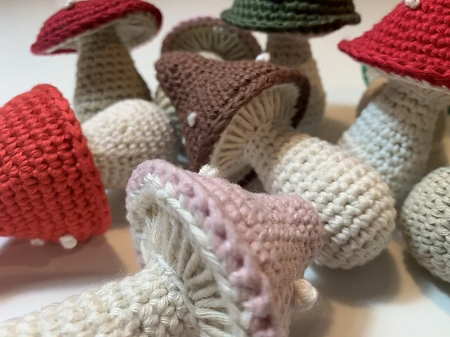 A group of Mushroom Amigurumi in various colors, including red, brown, pink, and green, are displayed closely together on a white surface.