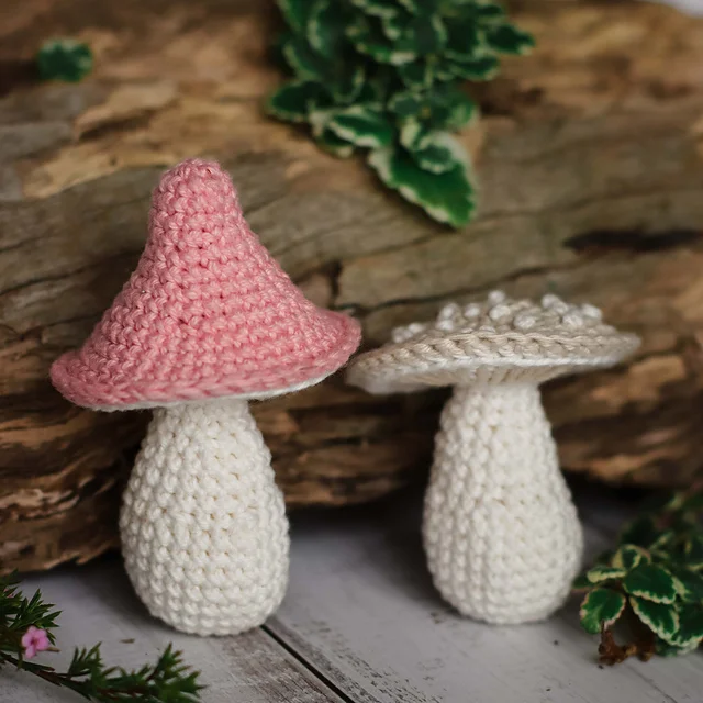Two crocheted Mushroom Amigurumi, one with a pink cap and one with a beige cap, are placed on a wooden surface with greenery in the background.