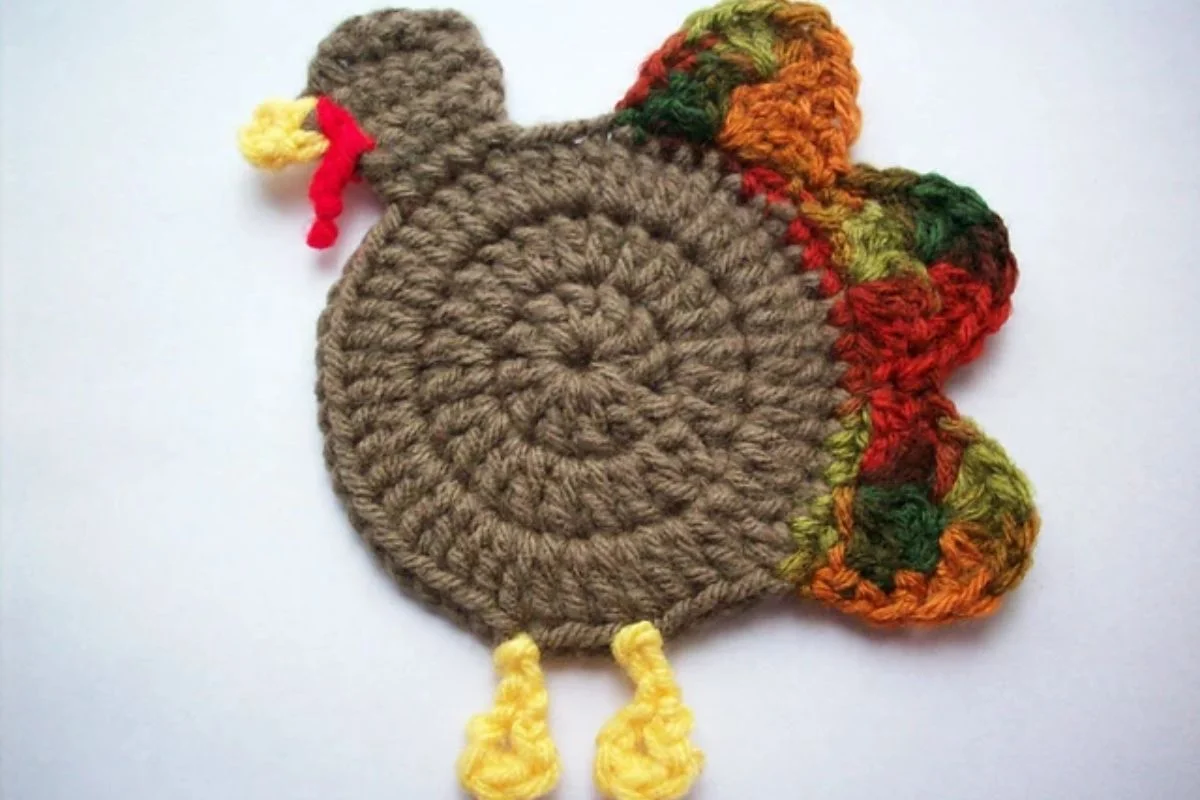 A crocheted turkey with a brown body, colorful feathers, and yellow legs against a white background.