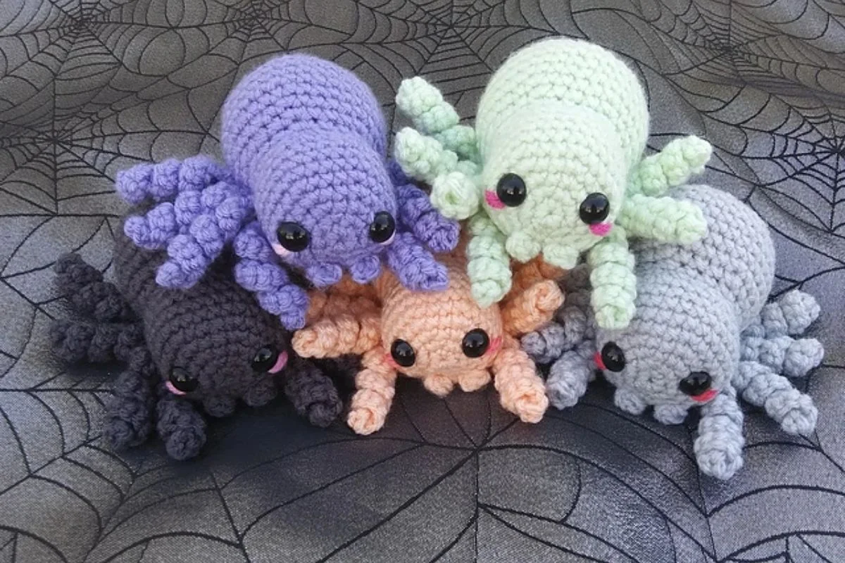 Amigurumi crocheted plush spiders in various colors, including purple, green, black, peach, and gray, are artfully arranged on a spiderweb-patterned background.