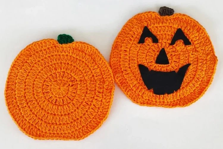 Two crocheted pumpkin coasters, one plain and the other with a jack-o'-lantern face, lie on a white surface.