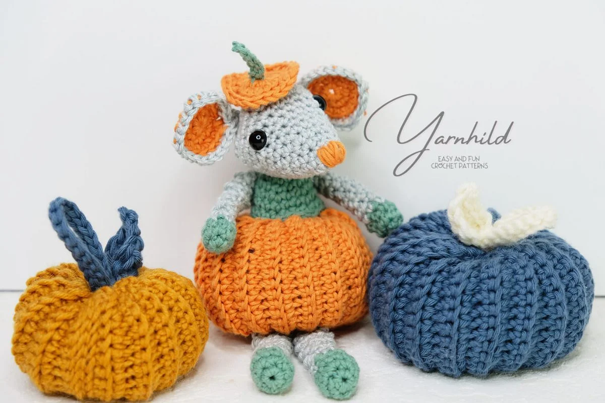 A crochet mouse in a festive pumpkin outfit sits charmingly between two crocheted pumpkins, one orange and one blue, capturing the whimsical spirit of Halloween.