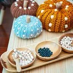 Three Crochet Pumpkin with Beads, featuring delicate white accents, are displayed behind wooden bowls containing crafting beads and crochet hooks on a wooden table.