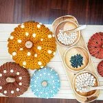 Four crochet pumpkin with beads, featuring white dots, are arranged alongside three wooden trays holding small beads on a light wooden surface.