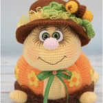A crocheted plush toy with a round face, big eyes, a brown hat with decorations, an orange outfit with yellow flowers, and a green ribbon bow around its neck—this charming creation is the perfect Autumn Turtle to brighten up your seasonal décor.