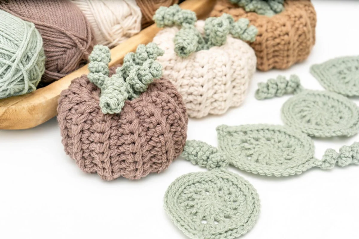 Crocheted pumpkins in brown and cream, adorned with intricate leaf designs, are displayed on a white surface. In the background, yarn balls in matching hues complement these charming crochet pumpkins perfectly.