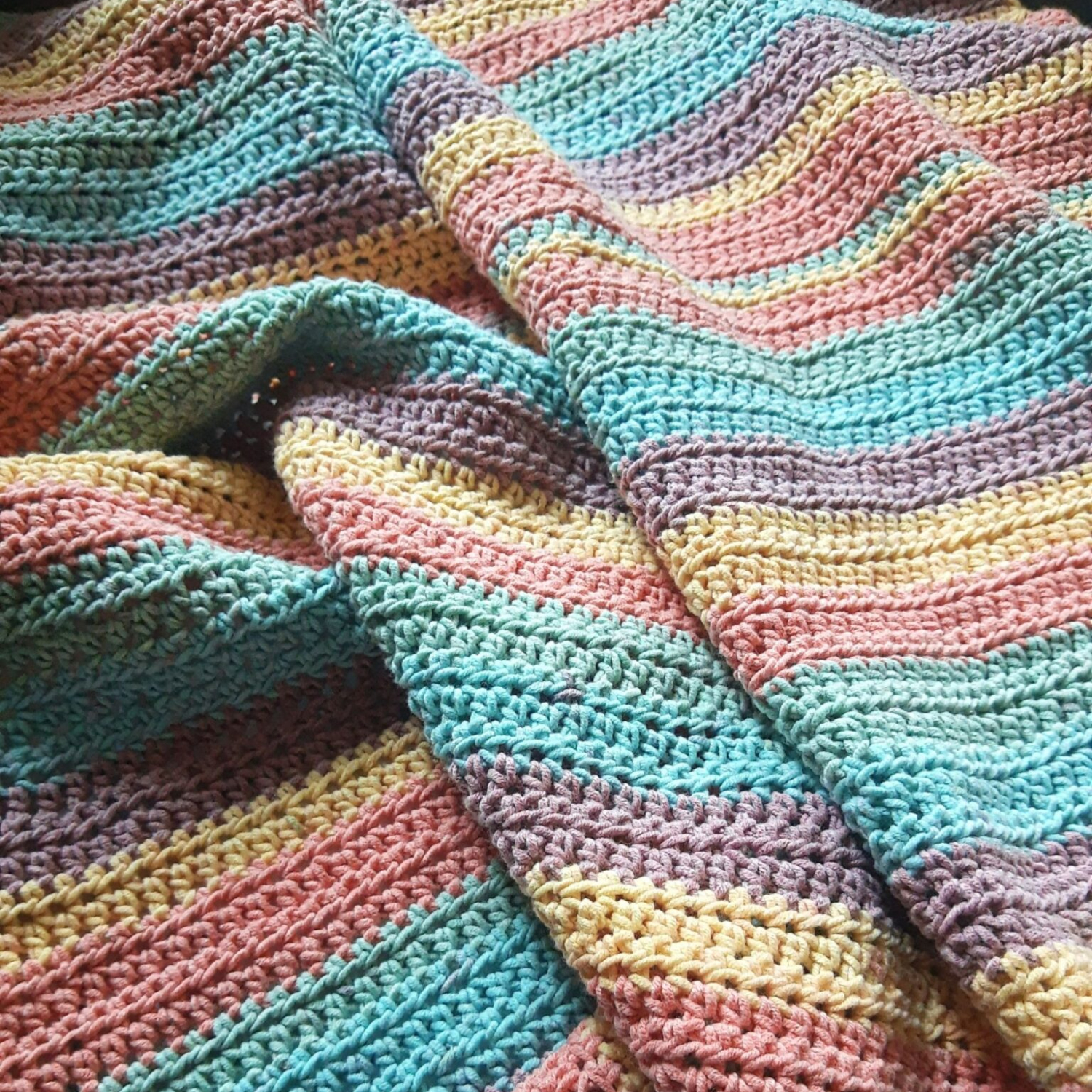 Crocheted blanket with colorful, wavy stripes in blue, yellow, pink, and green hues, spread out flat.